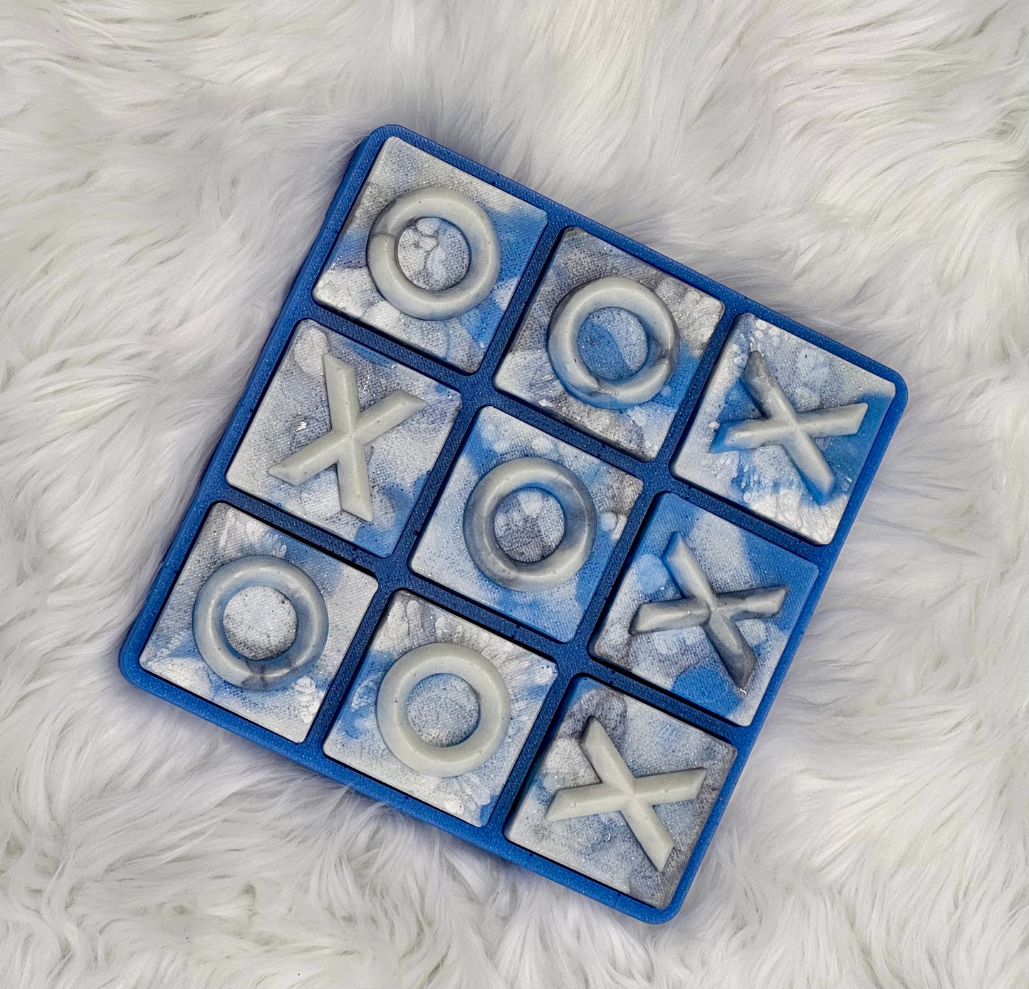 A blue square tic-tac-toe game board with marbled blue and white tiles, featuring white stone-like O and X symbols, photographed from above on a white faux-fur surface.