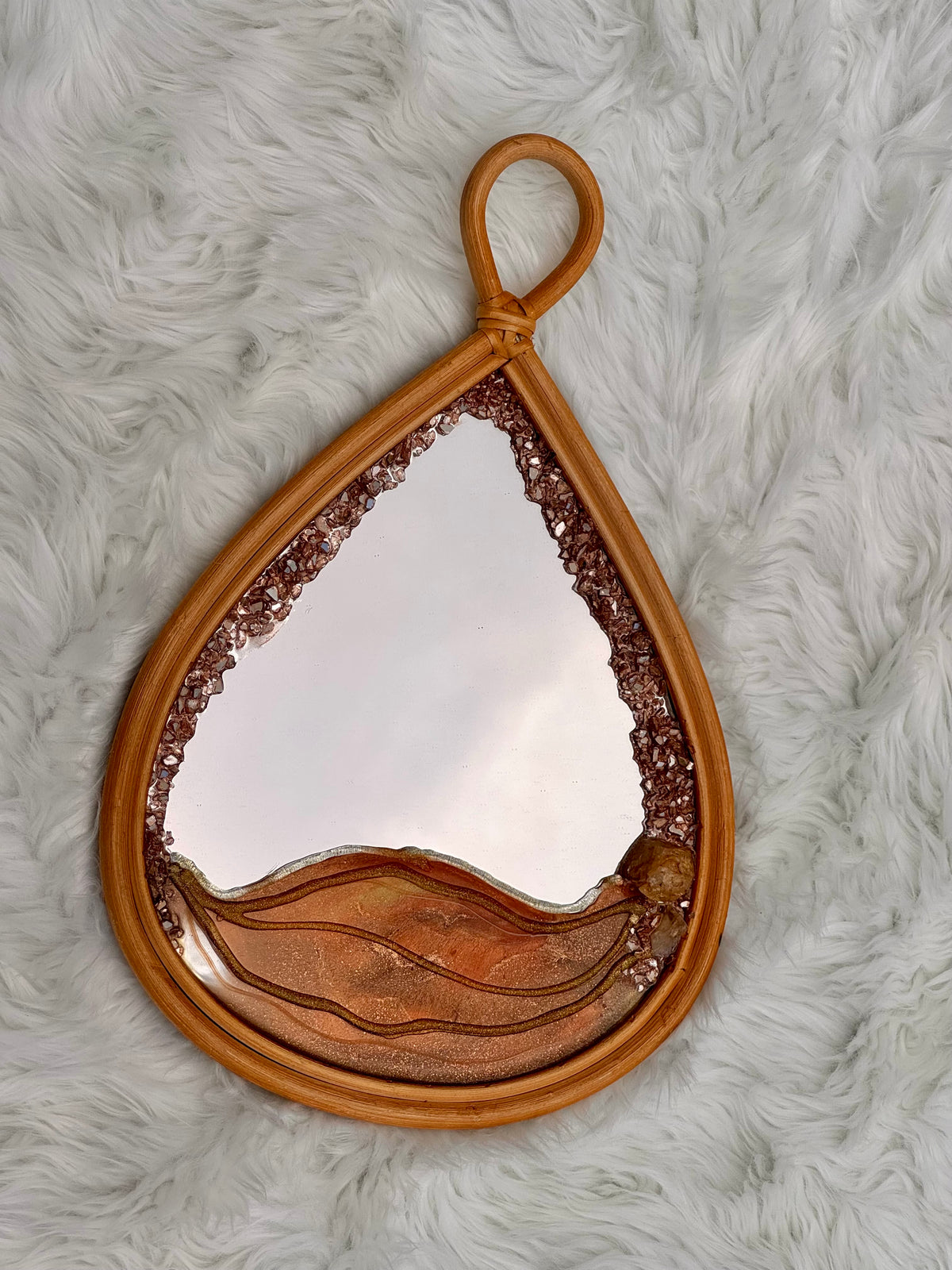 A teardrop-shaped decorative mirror with a warm brown frame, featuring textured metallic bronze wave details along the bottom and a beaded border around the reflective surface, displayed on a white faux fur background.