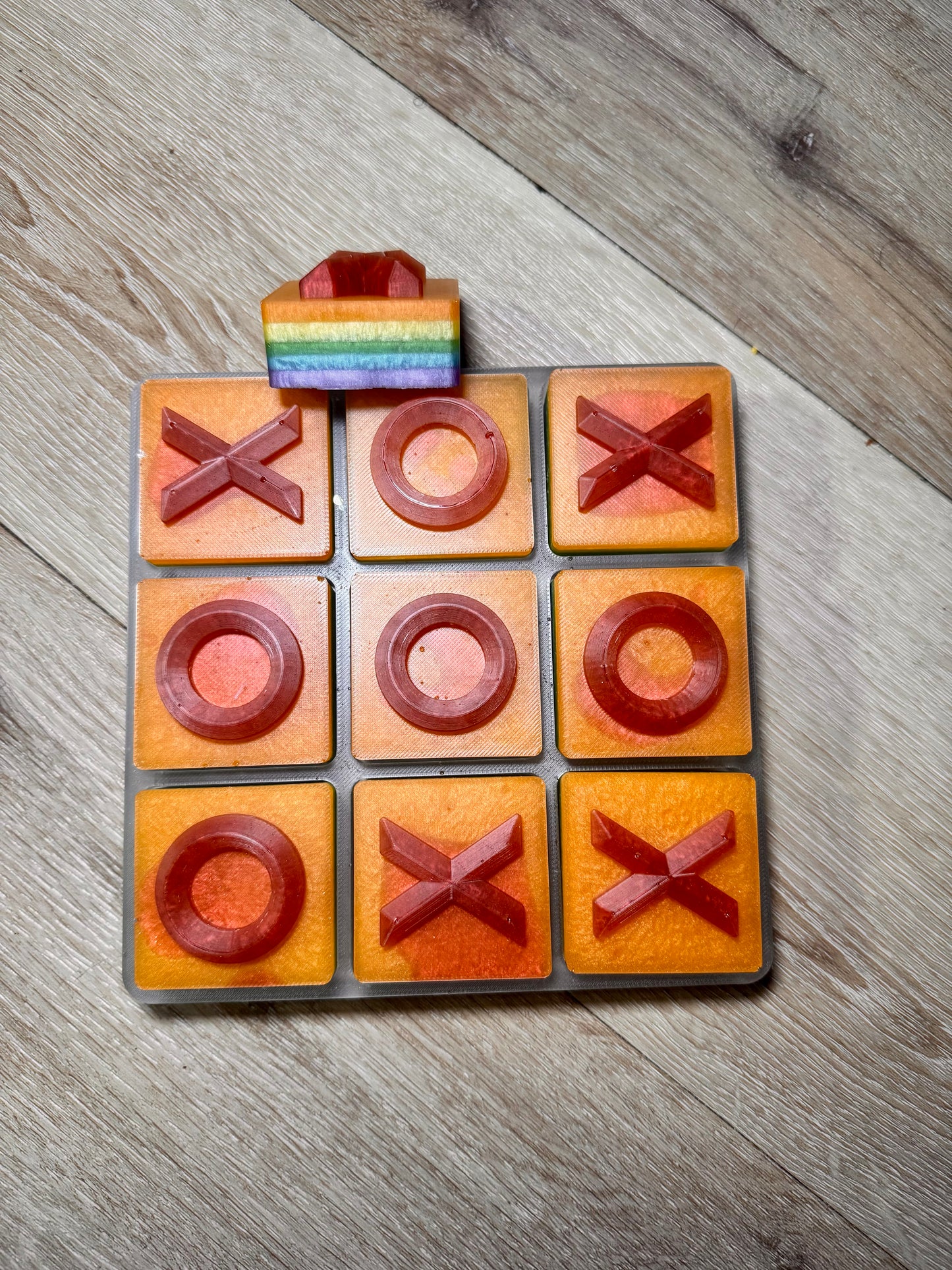 A 6.5x6.5 tic-tac-toe game board made from orange and red square tiles, with a small rainbow-striped block on top, photographed on a light wood floor.