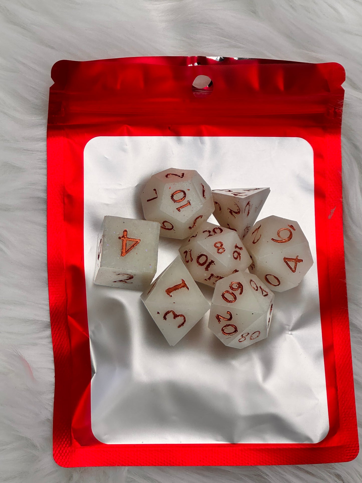 A set of translucent white polyhedral dice with red printed numbers, arranged on top of a red and white resealable pouch against a white faux fur background.