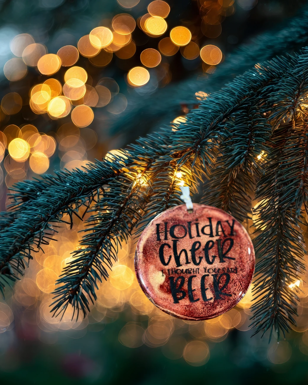 Decorative ornament with 'Holiday Cheer Through Your Beer' text on a Christmas tree branch with blurred lights in the background.