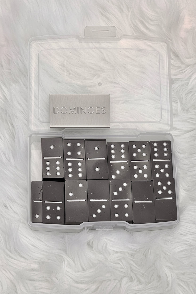 A set of black domino tiles with white pips in a clear plastic storage case, photographed from above on a white faux-fur surface.