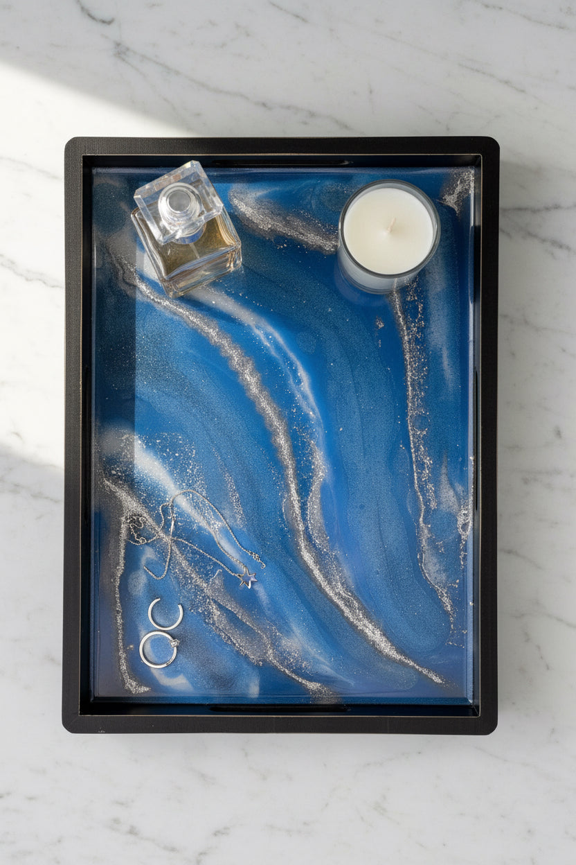 A square black-framed abstract art piece featuring a blue and white fluid marble pattern with embedded silver glitter, displayed on a white faux fur background.