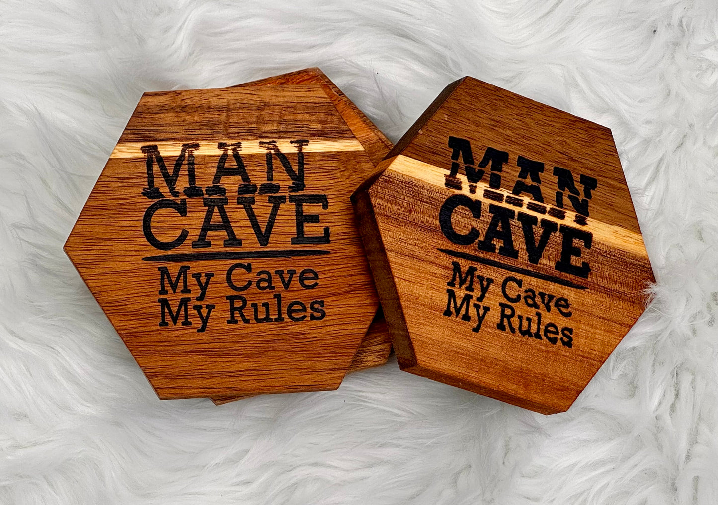 Four hexagonal wooden coasters with a natural wood grain finish, featuring the text 'MAN CAVE My Cave My Rules' engraved in black. They are displayed on a white faux fur background.