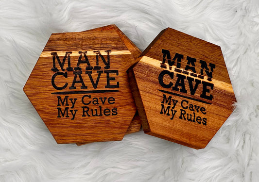 Four hexagonal wooden coasters with a natural wood grain finish, featuring the text 'MAN CAVE My Cave My Rules' engraved in black. They are displayed on a white faux fur background.