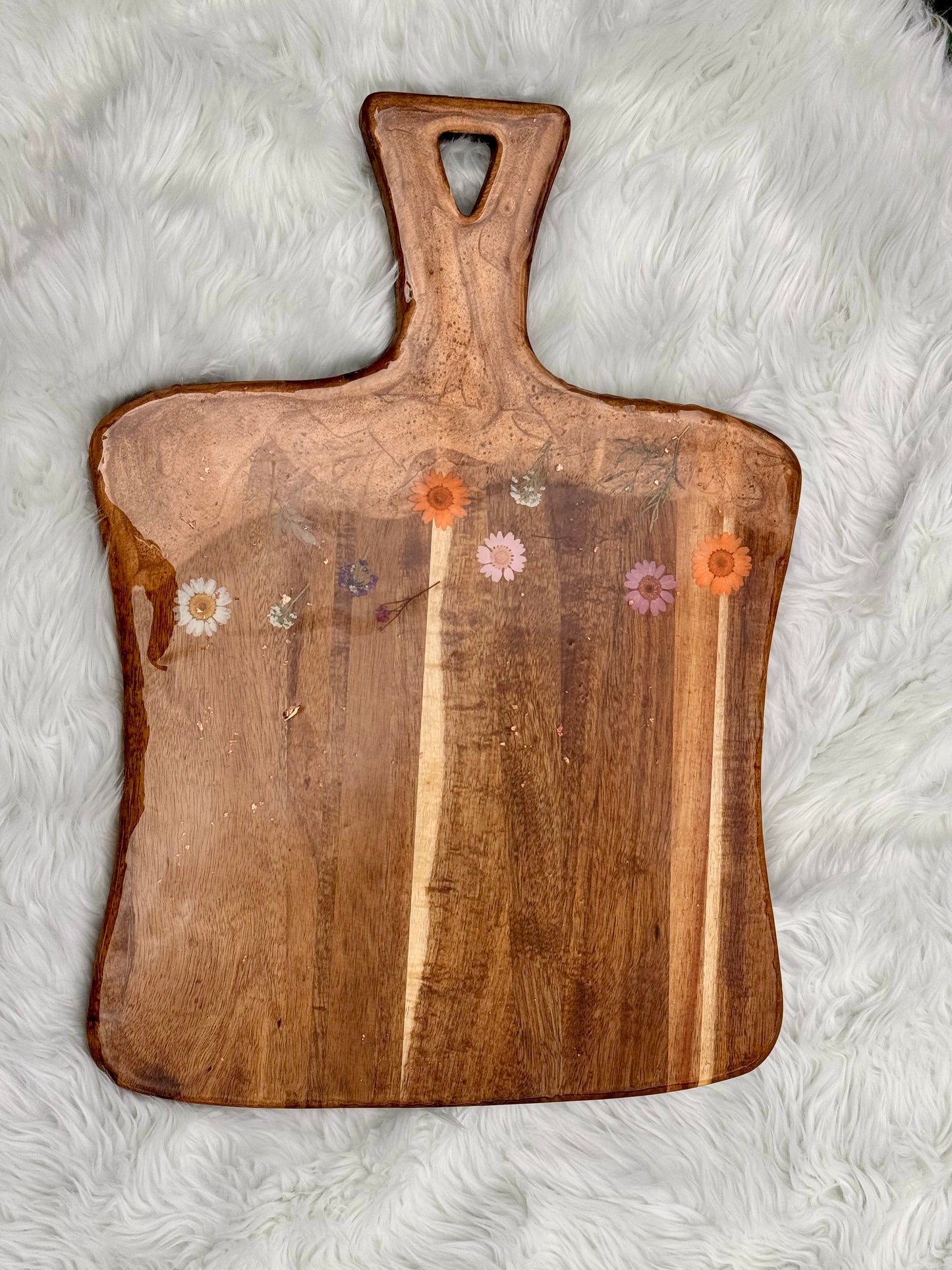 A rustic wooden serving board with a handle, featuring natural wood grain and small pressed flowers inlaid near the top, displayed on a white faux fur background.