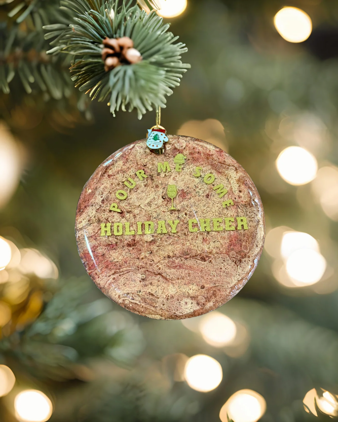 Round Christmas ornament with 'Pour Me Some Holiday Cheer' text on a tree with blurred lights in the background.
