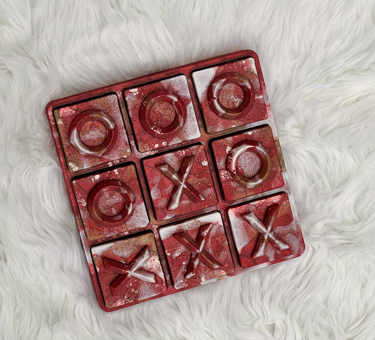 A square 3x3 tic-tac-toe game board with a distressed red and gold metallic finish, featuring embossed 'O' and 'X' symbols, placed on a white faux fur surface.
