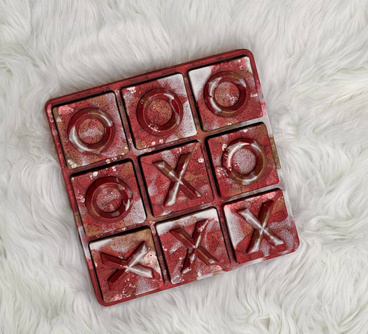 A square 3x3 tic-tac-toe game board with a distressed red and gold metallic finish, featuring embossed 'O' and 'X' symbols, placed on a white faux fur surface.