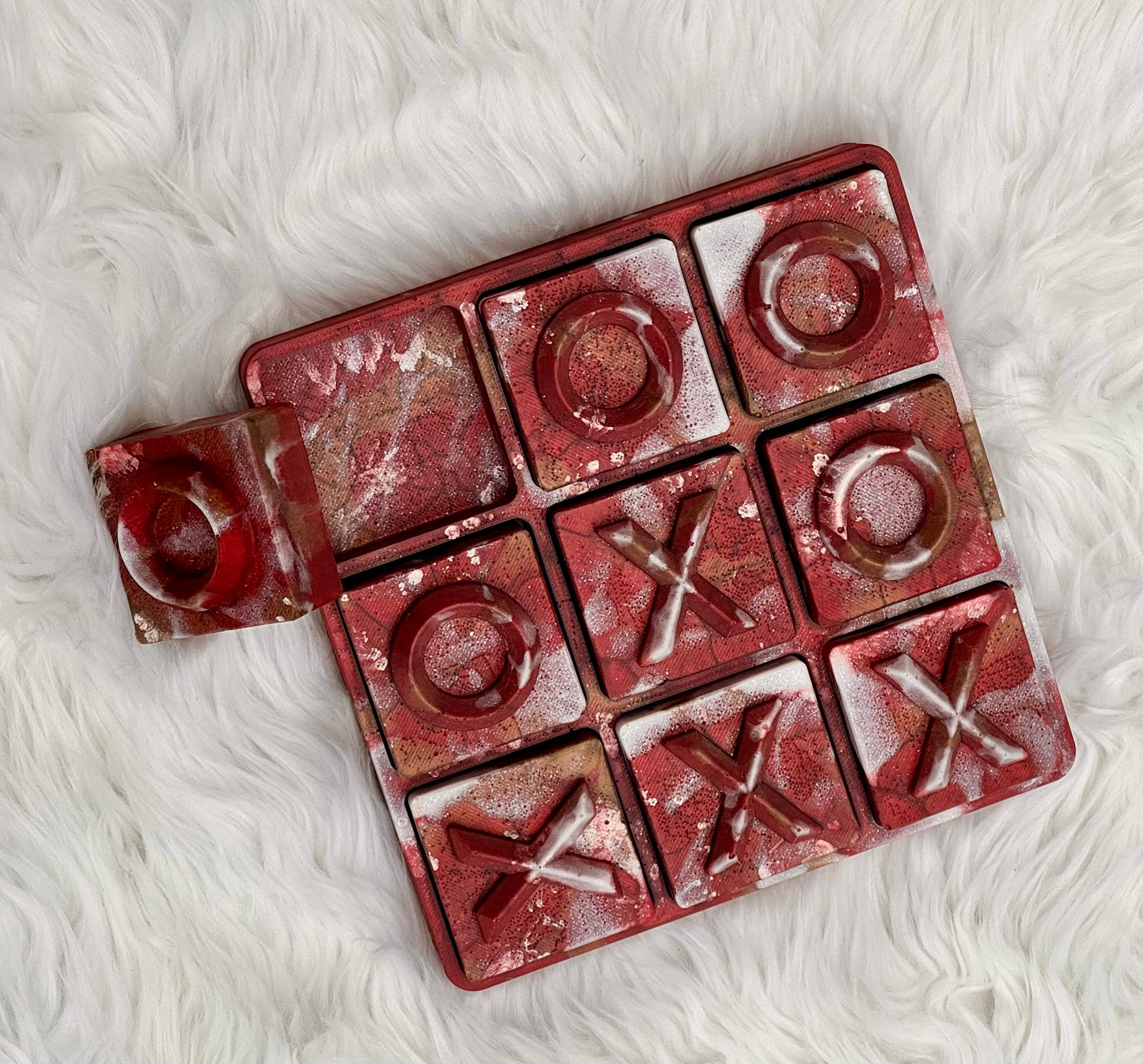 A square red tic-tac-toe game board with a small matching piece placed beside it, photographed from above on a white faux-fur surface.