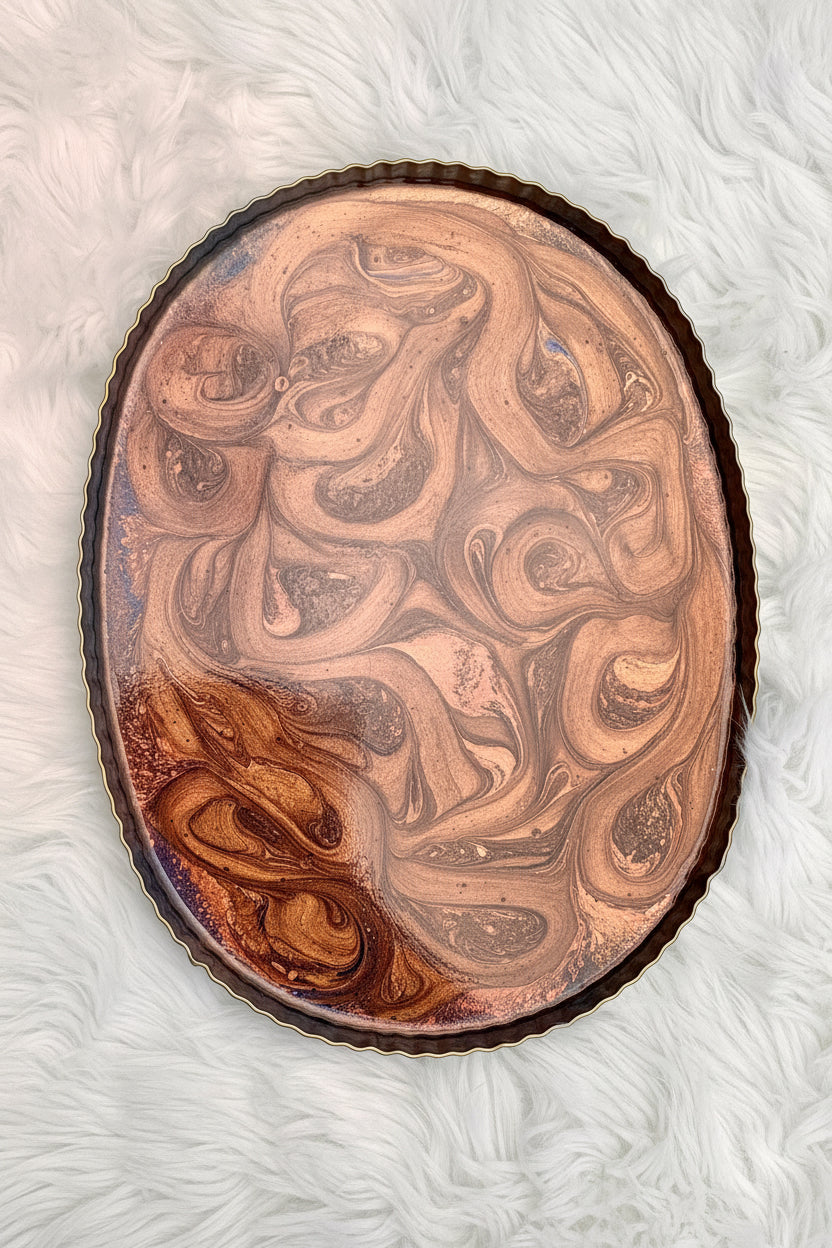 An oval decorative tray with a scalloped dark rim, featuring a glossy abstract swirl pattern in coppery bronze, brown, and beige tones, set against a white faux fur background.