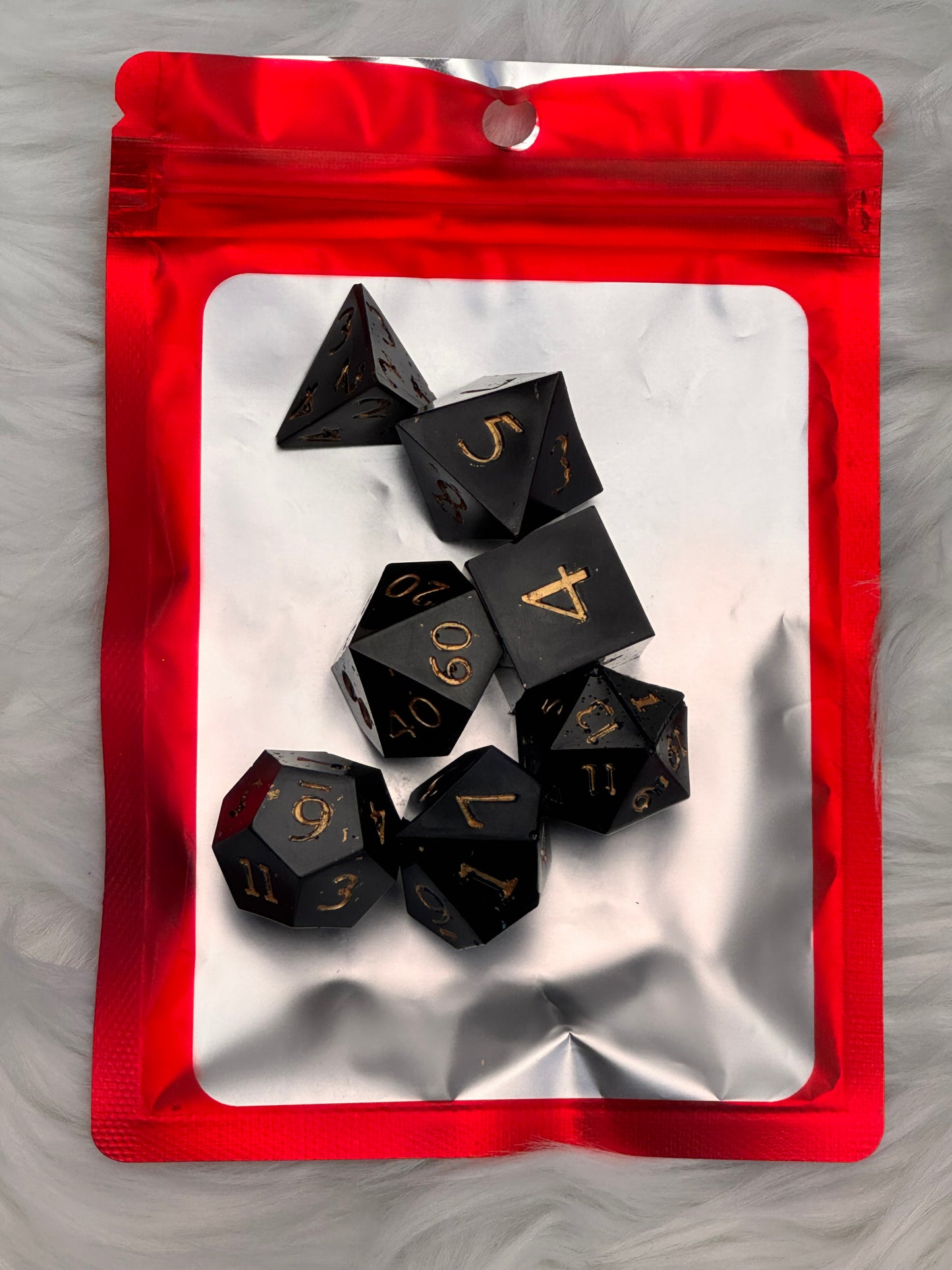 A set of black polyhedral gaming dice with gold numbers, arranged on a reflective silver tray inside a red resealable pouch, placed on a white faux fur surface.