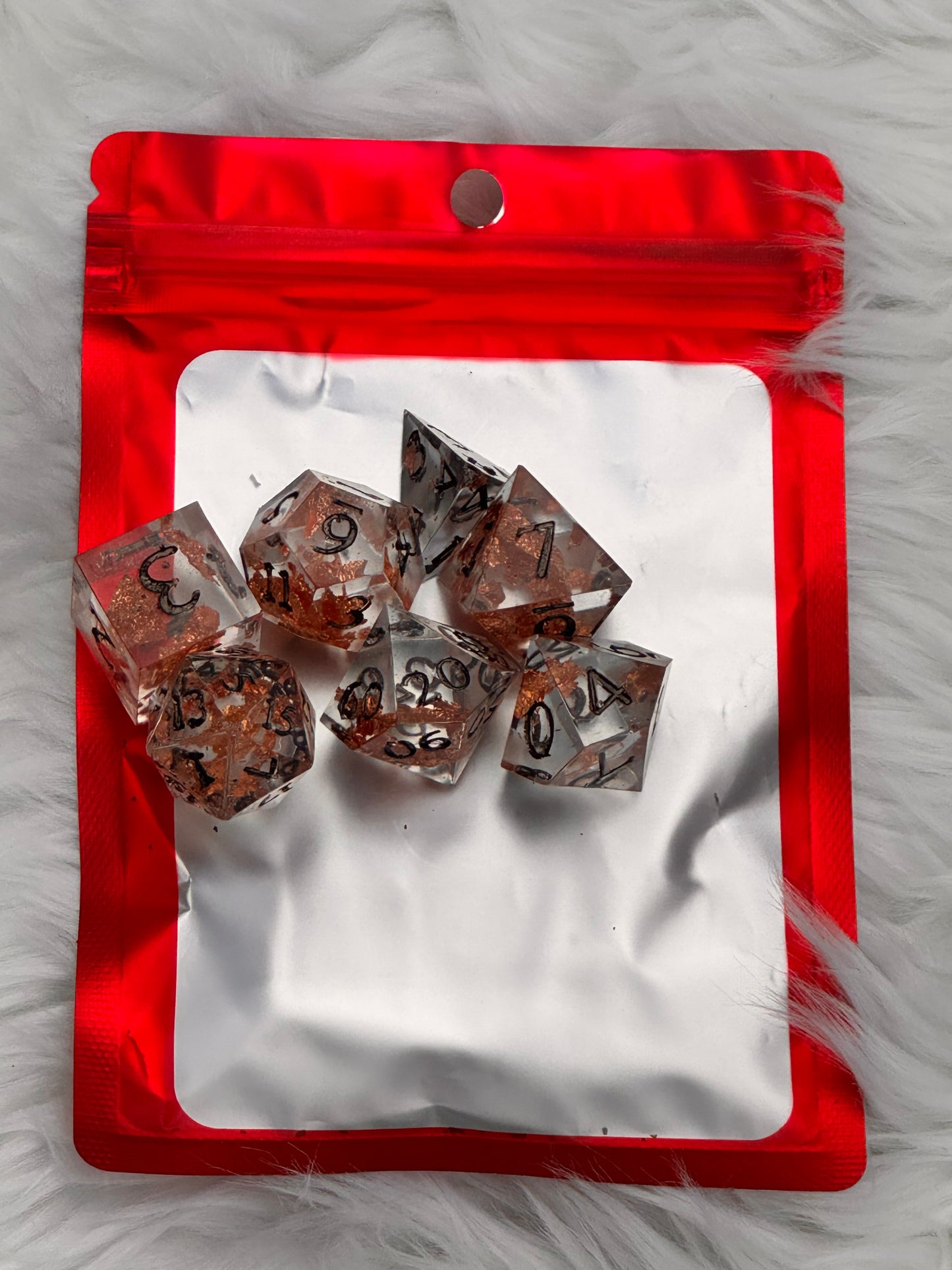 A set of clear acrylic polyhedral dice with brown and black speckled patterns, arranged on a red and white resealable pouch over a white faux fur background.