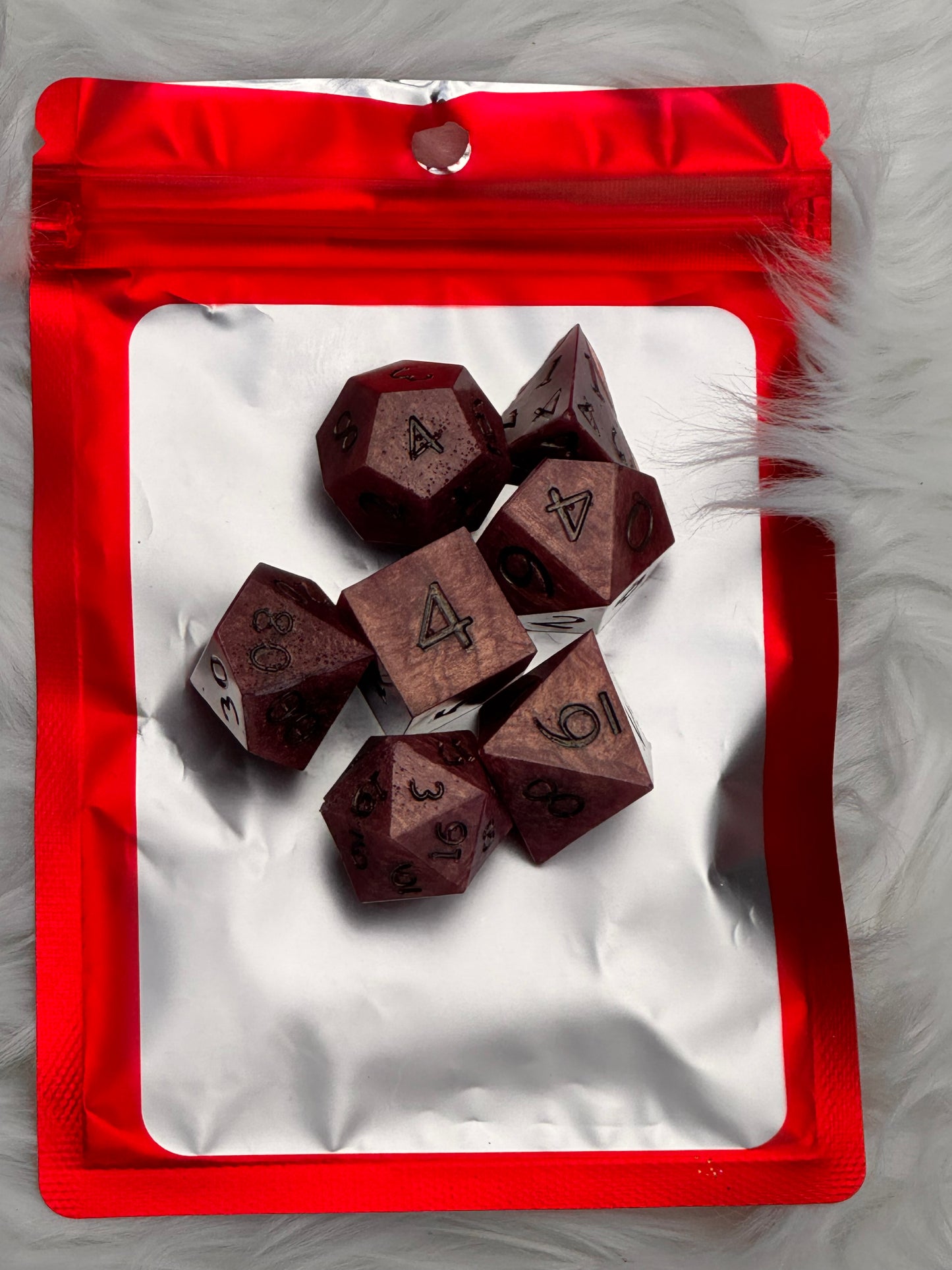 A set of dark red, wood-textured polyhedral gaming dice displayed on a white insert inside a red zippered pouch, set against a white faux fur background.
