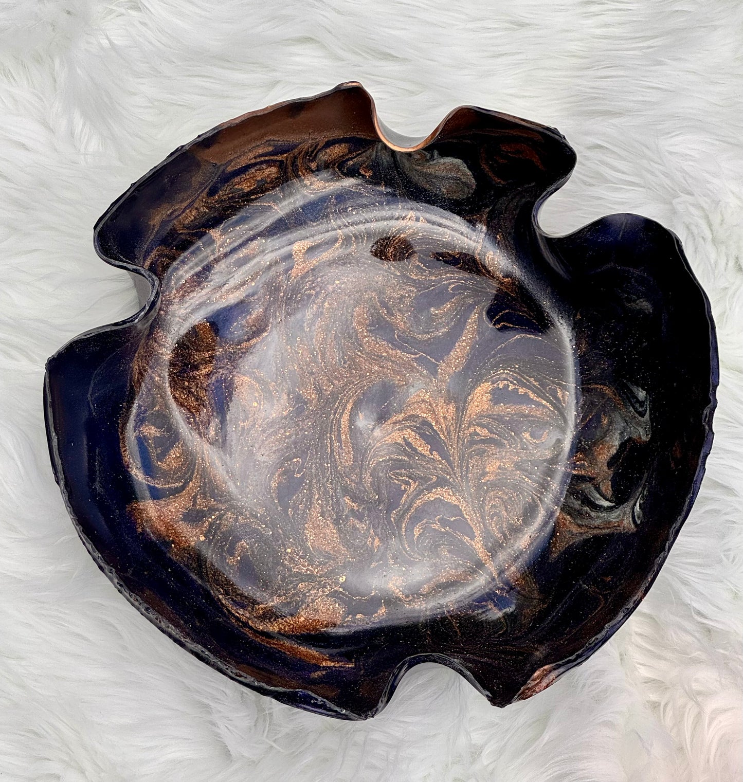 An organic-shaped resin bowl with a dark navy blue and bronze metallic swirl pattern, resting on a white faux fur surface.