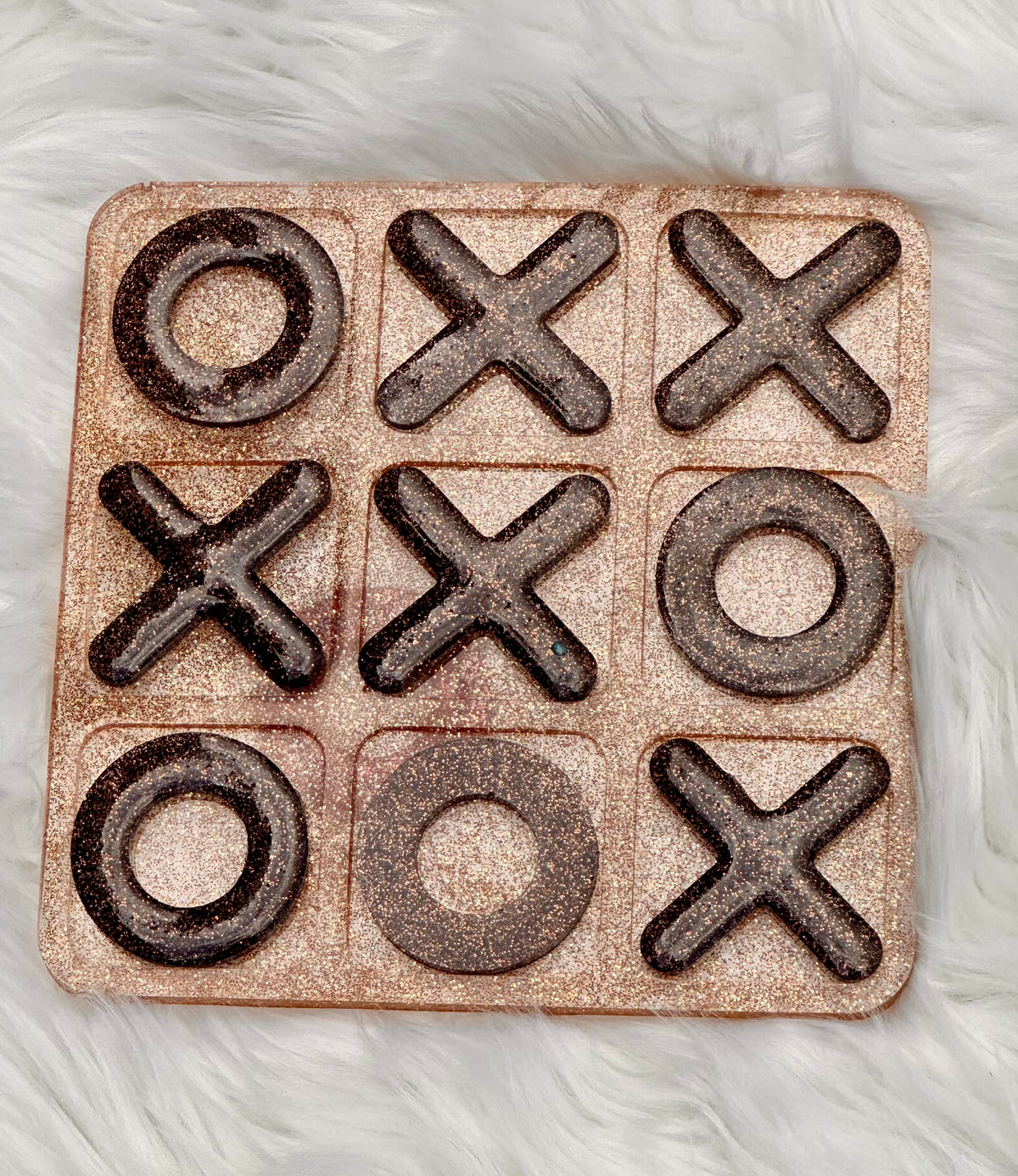 A square tic-tac-toe game board with a glittery rose gold finish, featuring dark brown embossed 'X' and 'O' symbols on a white faux fur background.