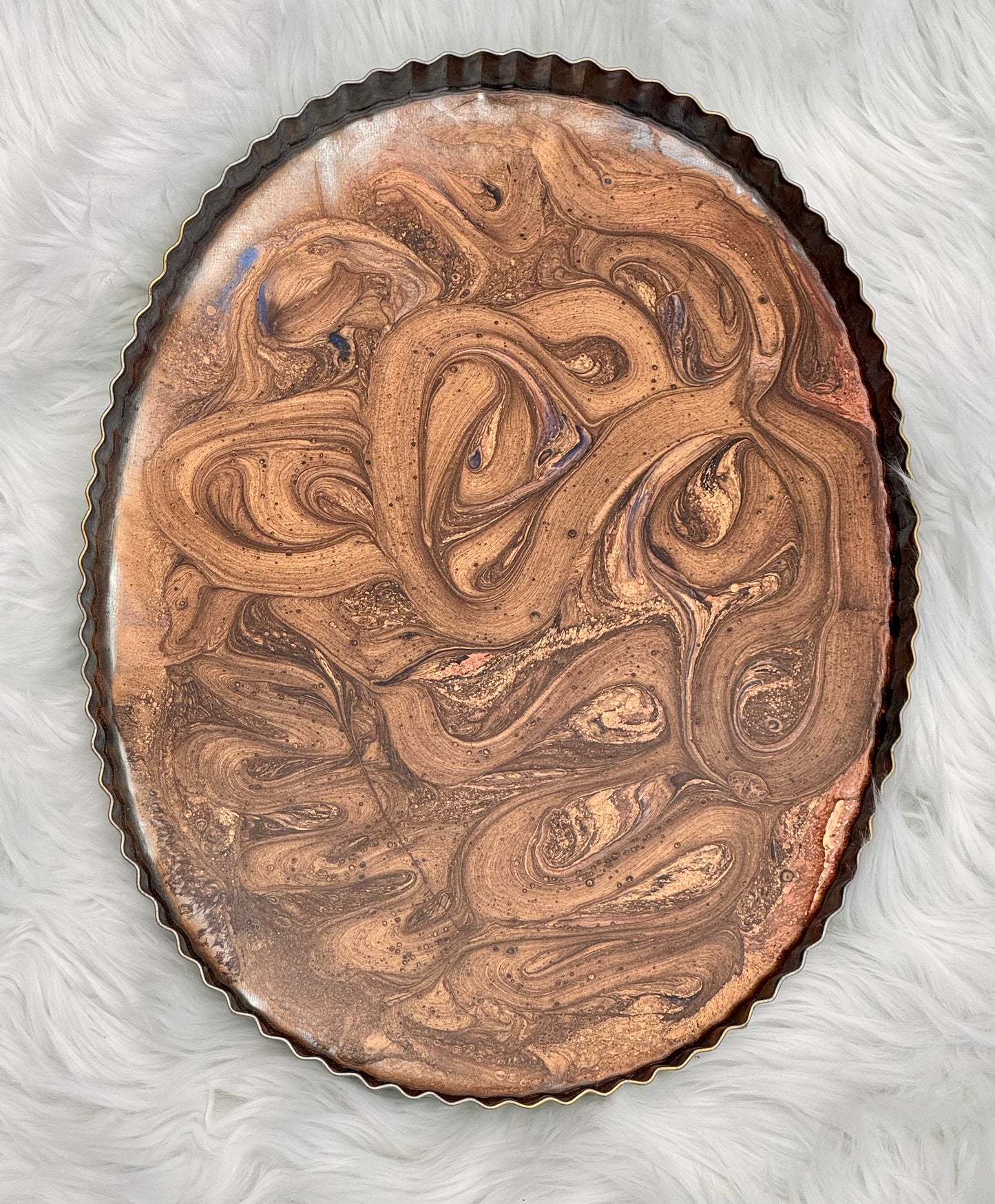 An oval-shaped metallic tray or medallion with a dark scalloped rim, featuring an abstract marbled swirl pattern in copper, bronze, and brown tones, set against a white faux fur background.