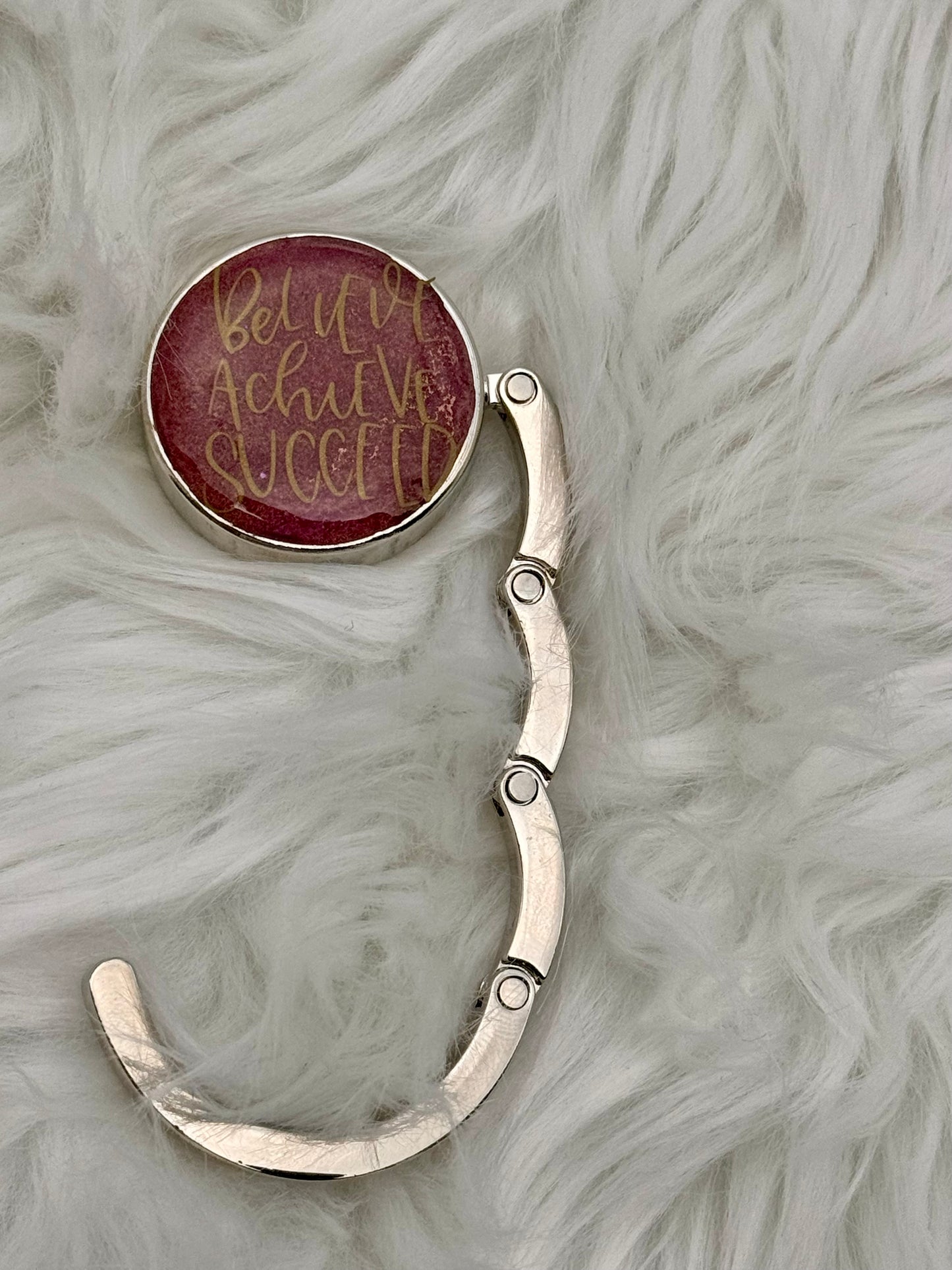 A silver-toned metal purse holder with a round burgundy cabochon featuring gold cursive text that reads 'Believe Achieve Succeed', displayed on a white faux fur background.