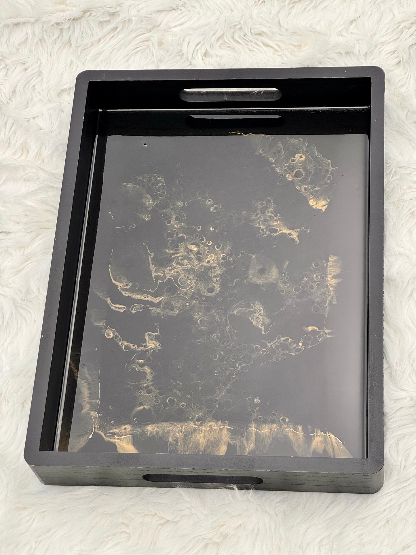 A rectangular black serving tray with cutout handles, featuring a glossy black and gold marbled pattern on the bottom surface, set against a white faux fur background.