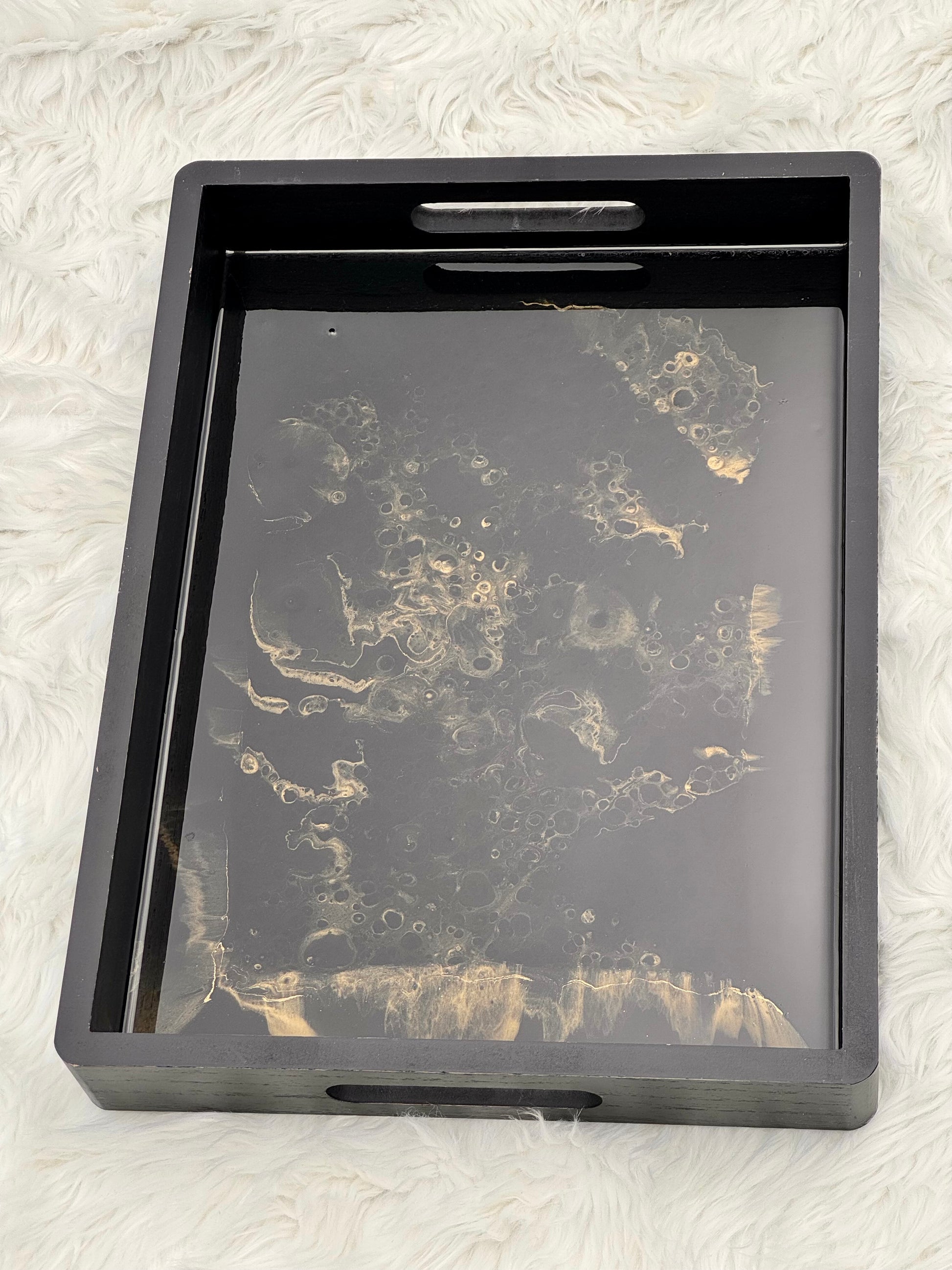 A rectangular black serving tray with cutout handles, featuring a glossy black and gold marbled pattern on the bottom surface, set against a white faux fur background.