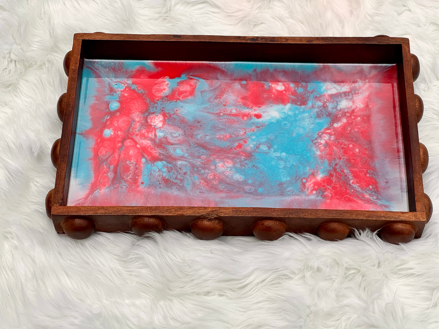 A rectangular wooden tray with a scalloped beaded edge and a red, blue, and white marbled resin insert, displayed on a white faux fur surface.