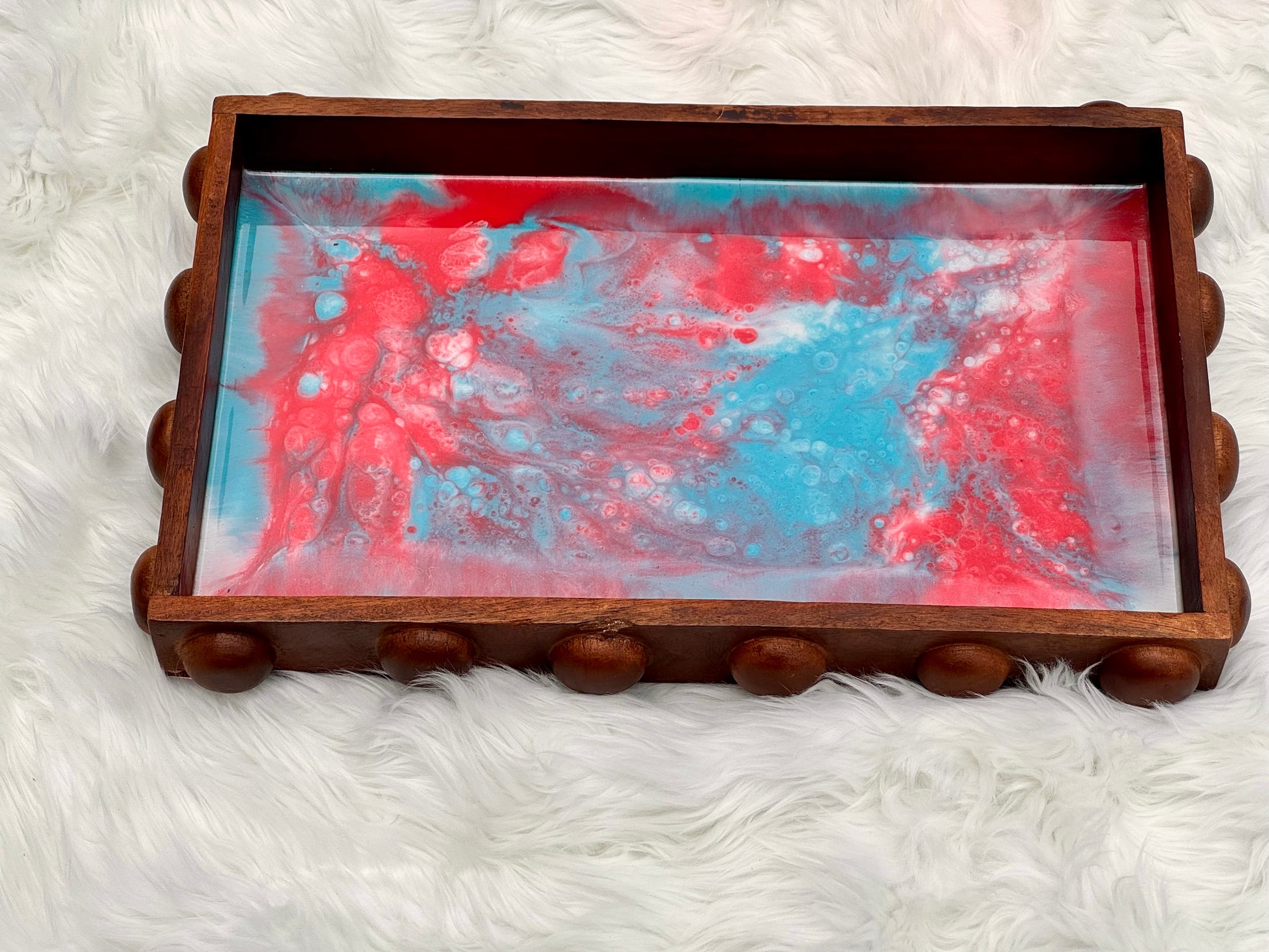 A rectangular wooden tray with a scalloped beaded edge and a red, blue, and white marbled resin insert, displayed on a white faux fur surface.
