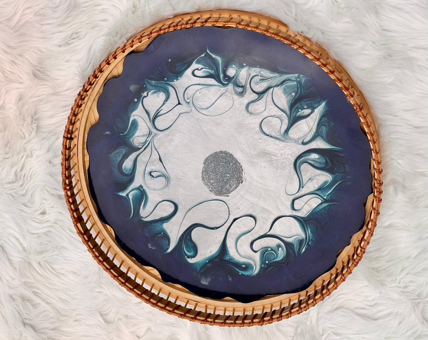 A round decorative tray with a woven natural wood rim and a dark blue-gray center featuring white and light blue marbled swirls, photographed from above on a white faux-fur surface.