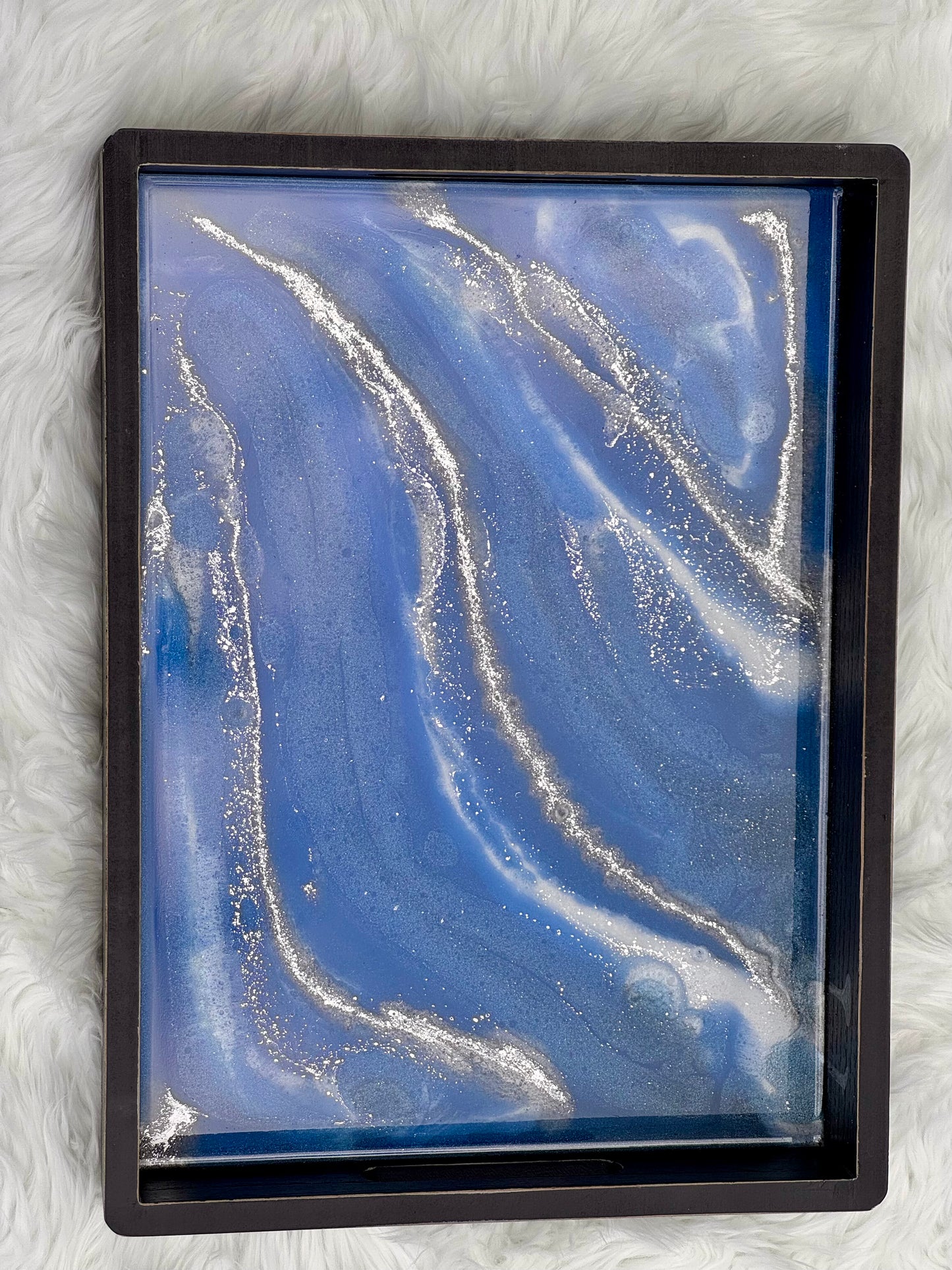 A square black-framed abstract art piece featuring a blue and white fluid marble pattern with embedded silver glitter, displayed on a white faux fur background.