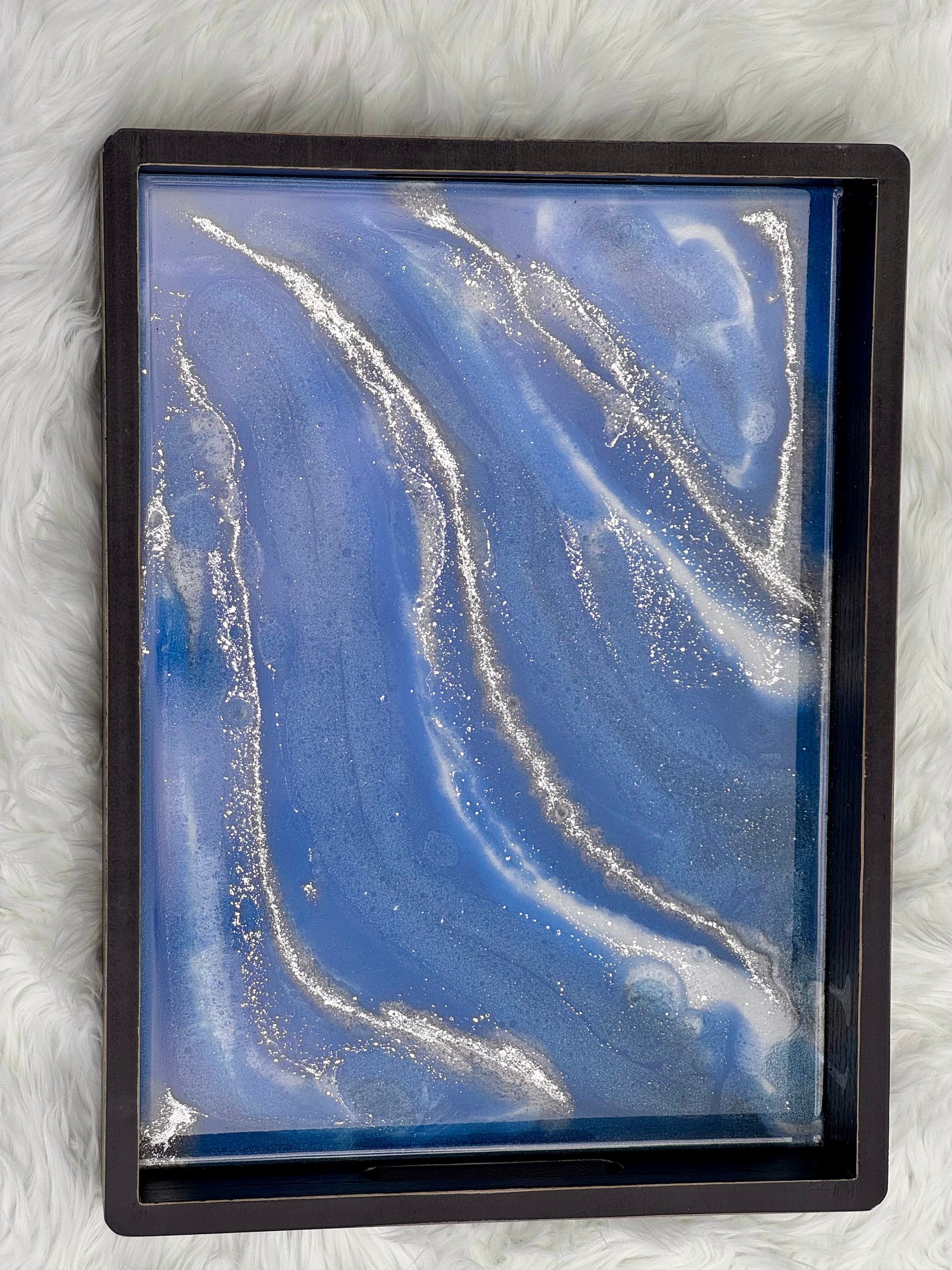 A square black-framed abstract art piece featuring a blue and white fluid marble pattern with embedded silver glitter, displayed on a white faux fur background.