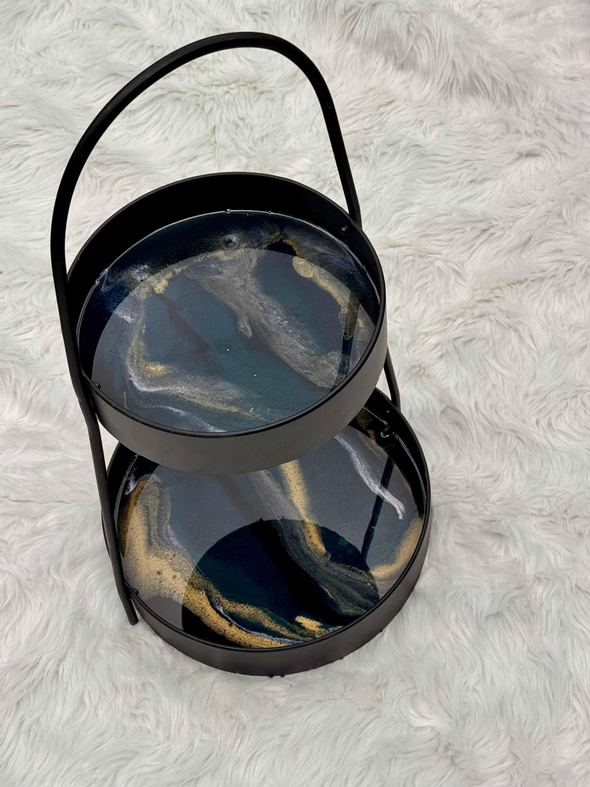 A two-tier round serving tray with a black metal frame and handle, featuring an emerald green and gold marbled resin finish, set on a white faux fur background.